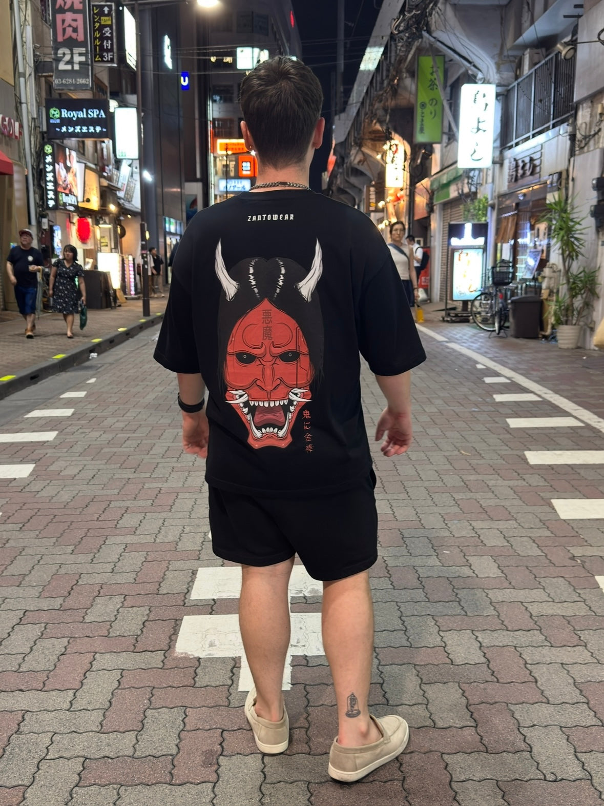 Person wearing a black t-shirt with a red oni mask design walking on a street at night.